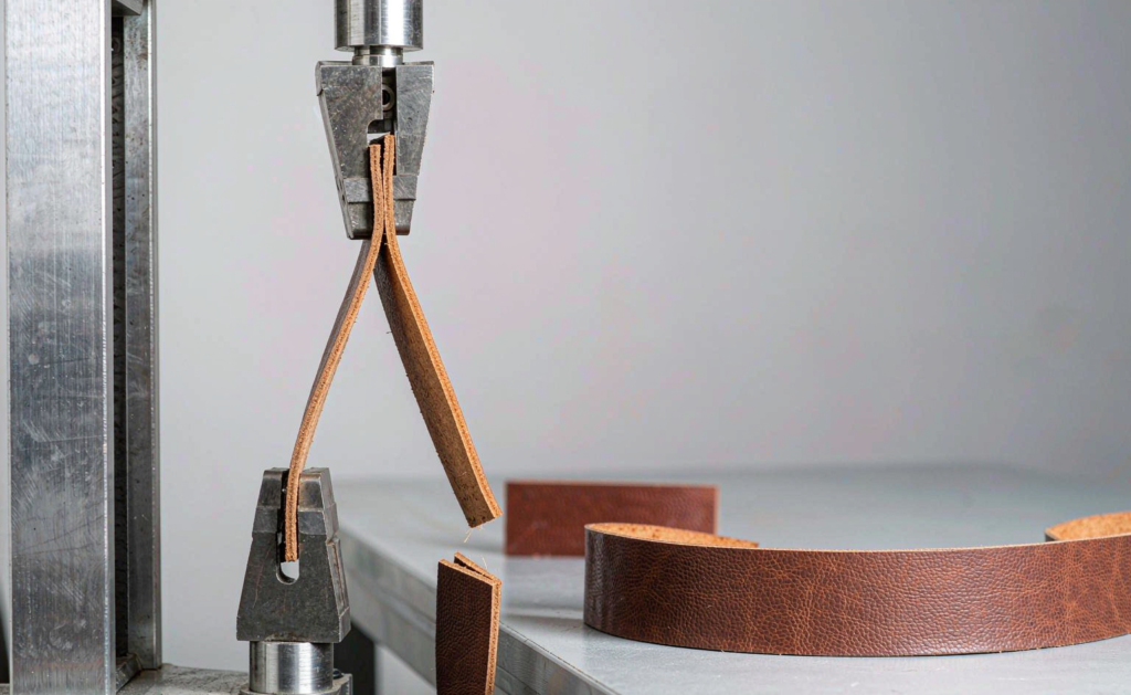 A tensile testing machine pulling a leather belt strap until it breaks, with a stretched sample and a comparison intact sample on a workbench